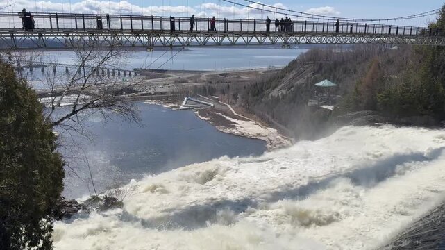 Wide panoramic view of a powerful waterfall cascading into a river basin. Pedestrians cross a suspension bridge high above the roaring water.