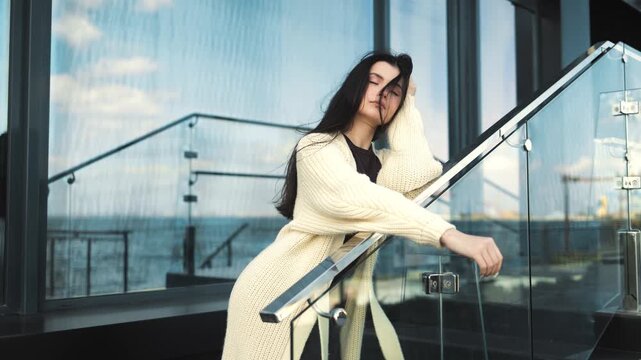 Student, girl, woman leans on a glass railing of a modern terrace, looking at the city view. Urban, lifestyle, fashion, elegance, city, morning, comfort, serenity, architecture, business, office