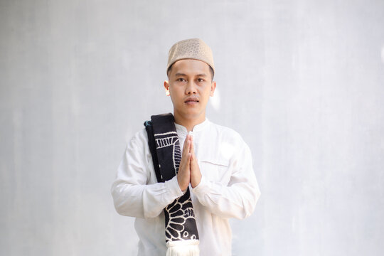 Portrait of a devout Muslim man in white koko shirt and peci, making a greeting gesture with a sarong on his shoulder against a white background