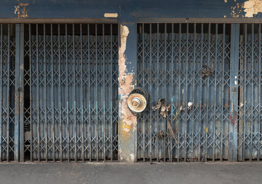A weathered, azure-hued steel folding gate traditionally known as Pratu Lek Yuet stands as a silent sentinel of classic Asian shophouse architecture. Characterized by vertical steel stanchions interla