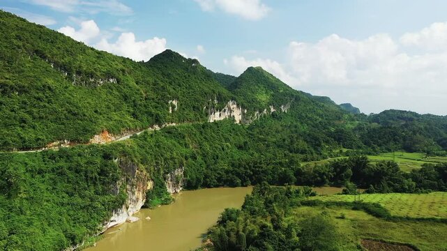 A scenic view of green mountains and a winding river near Lang Son, Vietnam, under a bright blue sky. The landscape features dense vegetation, rocky cliffs, and tranquil water.