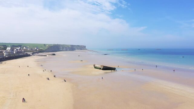 Wide sandy beach at Arromanches les Bains, Normandy, France, featuring scattered people and remnants of historic structures under a blue sky.