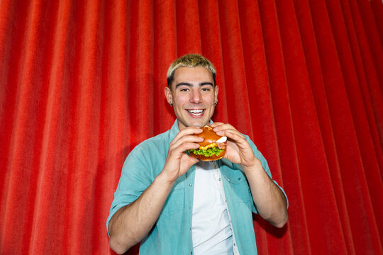 Smiling young man holding a burger before red curtain