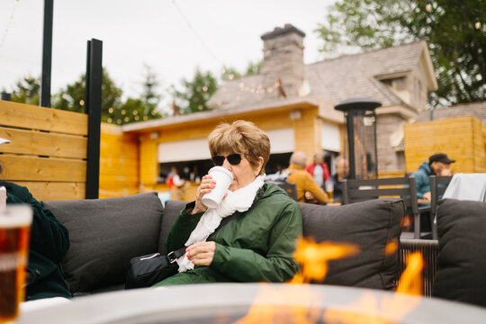 Elderly woman drinking coffee on outdoor patio near fire