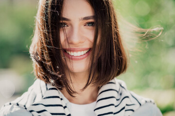 Obraz na płótnie Canvas Smiling young woman portrait outdoors in natural light with short brown hair and striped sweater, happy casual lifestyle close-up showing bright smile and fresh spring atmosphere
