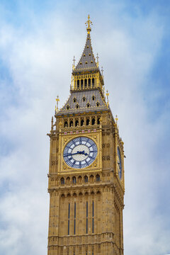 Close-up vertical shot of the Elizabeth Tower (Big Ben) featuring the iconic clock face and Gothic Revival architectural details against a cloudy blue sky, London, UK.