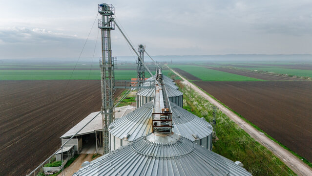 Aerial view of metallic silos standing tall against the backdrop of agricultural fields, where dark earth meets vibrant greenery, Sremska Mitrovica, Vojvodina, Serbia.
