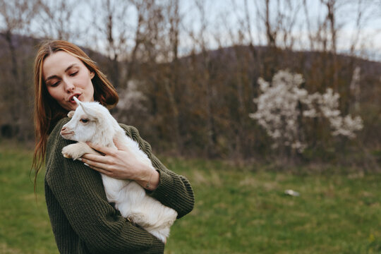 Caring woman holds a white goat kid outdoors in a park, sharing a tender moment. Lifestyle scene emphasizes nature, gentleness, and bonding, with soft natural light creating a calm, warm mood.