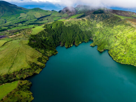 Aerial view of a stunning lake embraced by verdant hills and dense forests, where the water reflects the sky's tranquil blues, Flores, Azores, Portugal.