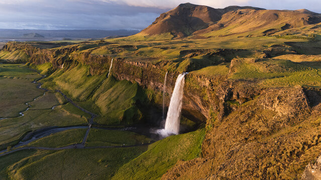 Aerial view of cascading Seljalandsfoss waterfall plunging from verdant cliffs at midnight sun into the Icelandic landscape, framed by rugged mountains, Seljalandsfoss, Iceland.