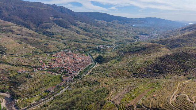 Aerial view of a compact town nestled in a valley, its terracotta roofs contrasting with the verdant slopes and rugged peaks, Cabezuela del Valle, Extremadura, Spain.