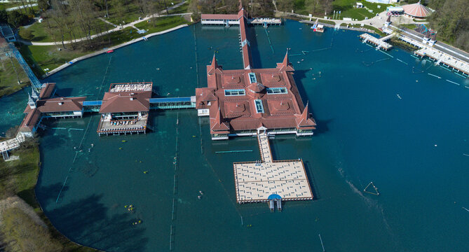 Aerial view of the vibrant turquoise waters embrace the iconic red-roofed buildings of Heviz Lake, creating a striking contrast against the surrounding lush greenery, Heviz, Zala, Hungary.