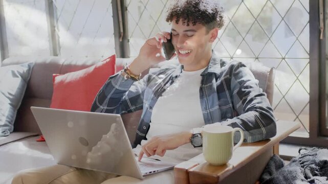 Adult man answering phone call while typing on laptop for remote work, smiling by window