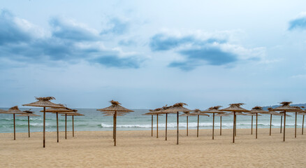 Serene sandy beach with straw umbrellas under a cloudy blue sky. Peaceful seaside landscape, perfect for travel and vacation themes, highlighting a quiet coastal atmosphere © John_Doo78