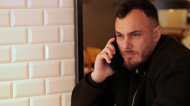 A medium shot of a young man with a beard, wearing a black jacket, engaged in
a phone call while sitting in a modern cafe. A glass mug of herbal tea with a
visible tea bag is on the wooden table 