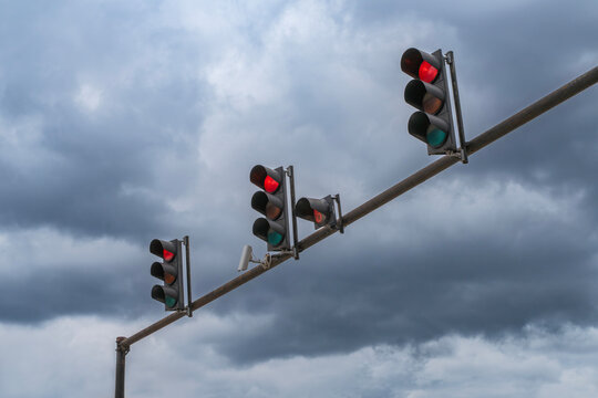 Three traffic lights showing red against a dramatic, dark cloudy sky. A row of signal lamps and a security camera mounted on a metal pole over a city road