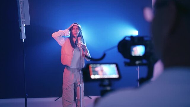 Backstage view of a cameraman filming a music video. A young female singer, wearing a baseball cap, is rapping into a microphone in a studio