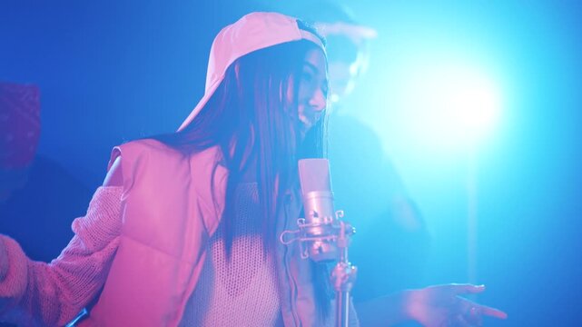 Energetic female artist with long dark hair singing into a condenser microphone. The singer performs passionately in a studio with blue lights