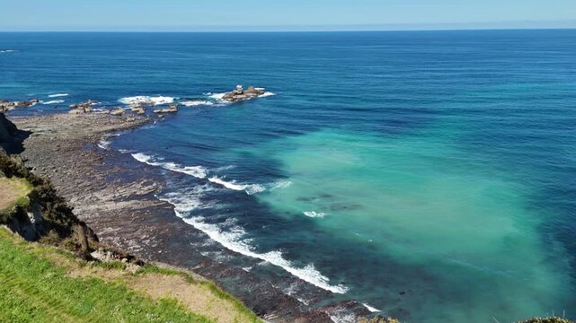 Panoramic drone view capturing the rugged Asturian coastline with green cliffs, a rocky beach, and the blue Atlantic Ocean under a clear sky in Northern Spain