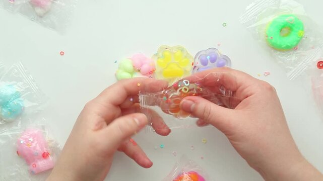 Top view of hands on a white table gathering pastel squishy charms, opening clear wrappers, lifting and squeezing the toys one by one, then stretching the transparent material in sensory play.