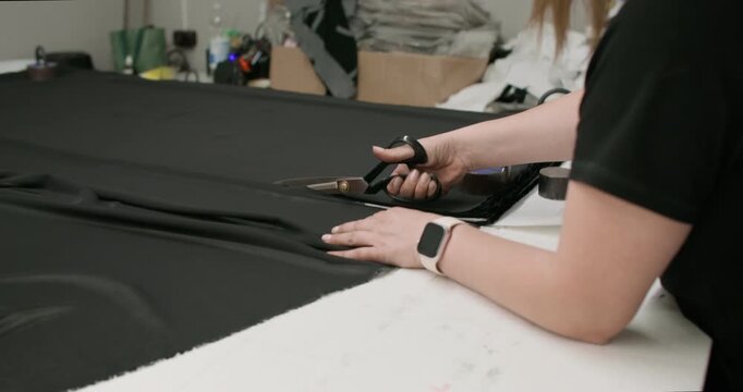 Sewing workshop. Clothing production. Female seamstress pressing black fabric to table with weights cutting scissors piece from roll.