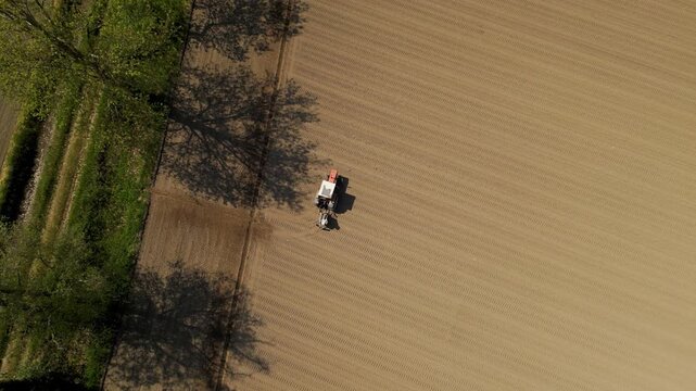 Cultivating the Earth: An aerial perspective captures a lone tractor diligently tilling a vast agricultural field, amidst the serene embrace of budding trees. 