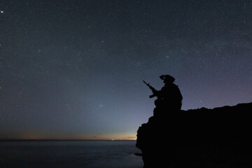 Silhouette of a tactical special forces soldier with night vision goggles sitting on a cliff under a vast starry sky © Dmitri