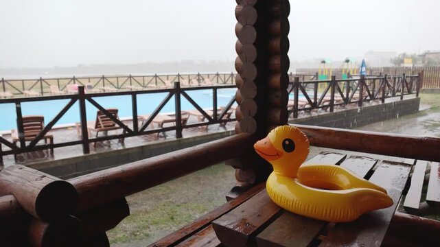 An inflatable duck lies on wooden table in gazebo at recreation center against the backdrop of pool covered with rain