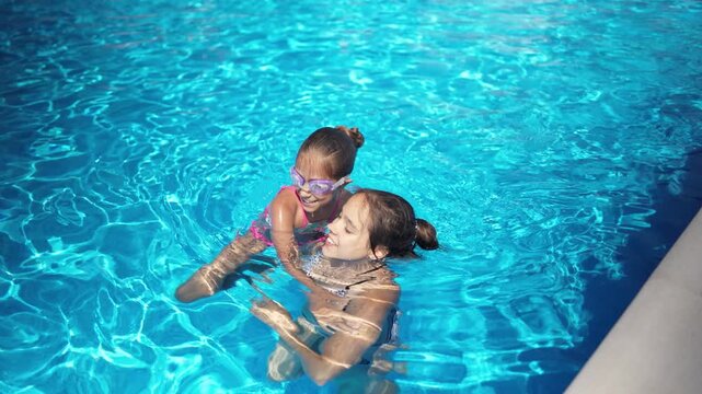 Two girls in bright bikinis are playing together in a pool with blue water on a summer day on a long-awaited vacation