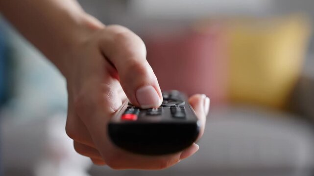 This shows a close-up shot of a person's hand holding a TV remote control. The background is blurred, but it appears to be a living room with a couch.