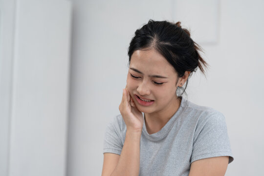 Young Asian woman suffering from toothache, touching cheek with pain, dental problem, sensitivity or cavity, oral health issue and healthcare concept in home environment