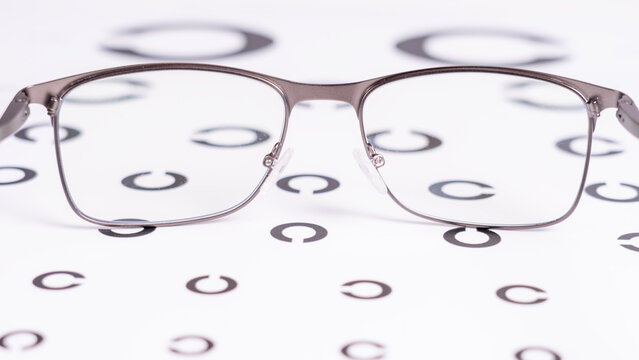 Modern metallic eyeglasses rest on a white eye chart. Black circles of varying sizes form the standard visual acuity test. Thin metal frame with clear lenses highlights the eyewear's design
