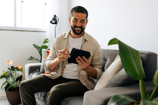 Man sitting on sofa at home using tablet, browsing, reading or communicating in a bright living room environment with natural light.