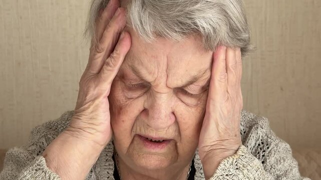 An elderly grandmother dressed in a knitted sweater is sitting in a room, sad face, headache, holding her hands on her temples leaning on the table, looking away with her eyes. Sadness. Close-up.