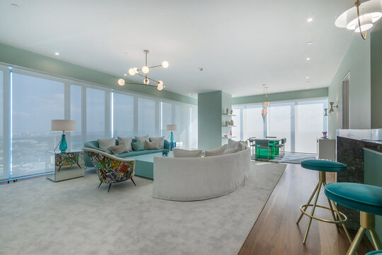 A spacious living room features large windows that let in sunlight. Colorful furniture and a modern style create an open and inviting space. The view outside shows a busy city skyline