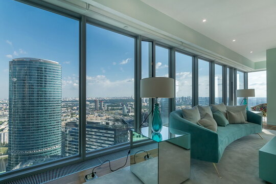 A view from a high rise building shows a large city with many buildings. There are modern furniture and lamps near the windows. It is bright outside with clear blue skies