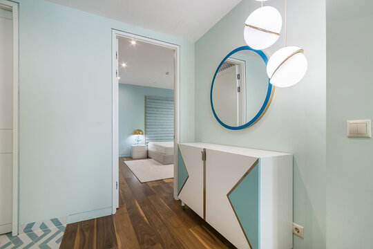A hallway shows light blue walls and wooden flooring. It leads to a bedroom with a round mirror and a lighting fixture. A cabinet with a simple design sits against the wall