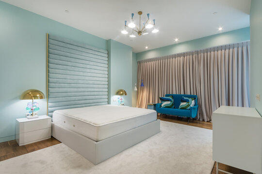 The room shows a bed without a mattress, blue walls, a green sofa, and curtains. Two tables with lamps are beside the bed. Lights are on, creating a warm space