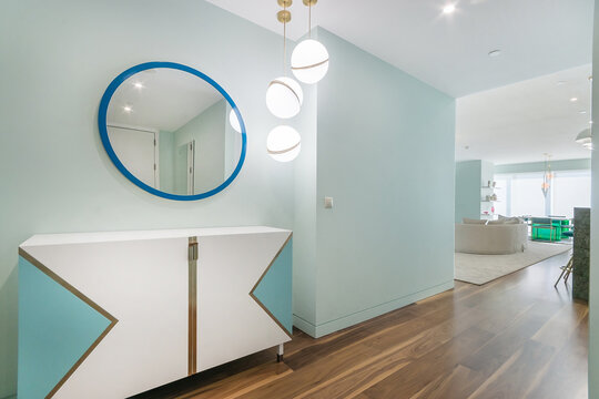 A hallway features a round mirror above a white cabinet with blue accents. Modern light fixtures hang from the ceiling. The entrance leads to a light-filled living area