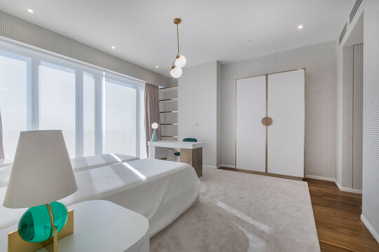 In a bright room, large windows let in sunlight and show a clean design. There is a bed, a desk, and a lamp. The space feels open and organized, promoting functionality and comfort