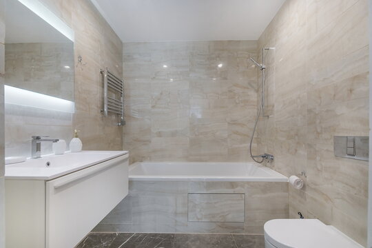 A bright bathroom features a bathtub and a shower. The walls and floor have beige tiles. The room has a mirror and a towel heater. Natural light comes through the space