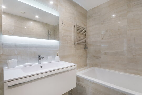 A bathroom features a white bathtub, a sink with a mirror above, and towel racks. Natural light brightens the space. The walls and floor have a light stone finish