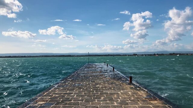 A beautiful view of a long stone pier extending into the choppy turquoise sea with waves splashing over it under a bright blue sky with fluffy white clouds.

