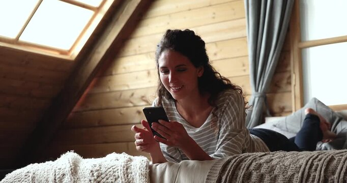 Smiling adult woman lying on bed using smartphone, checking message, chatting with friend, enjoy online communication. Personal leisure, digital lifestyle, relaxation at home, mobile application usage