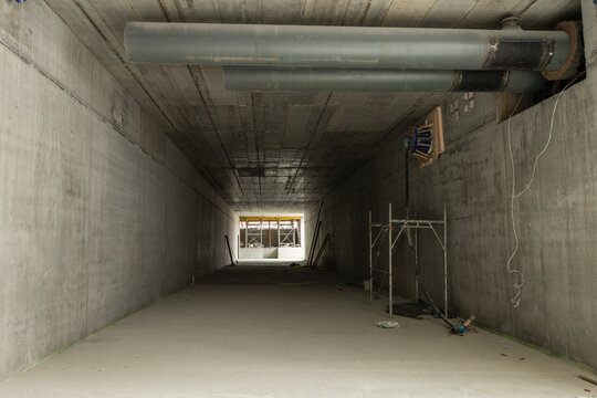 Wide-angle view inside a raw, unfinished concrete pedestrian underpass.. Raw concrete surfaces, exposed horizontal pipes with valves and a gauge panel, scaffolding, and a large opening with strong