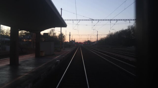 Railway station view from moving train at dusk with glowing signal lights overhead wires and calm evening atmosphere guiding tracks into horizon