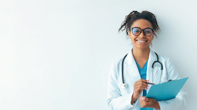 A young female doctor stands by a white wall in an office. She smiles while holding a clipboard. She wears a white coat and has glasses on. Her stethoscope hangs around her neck.