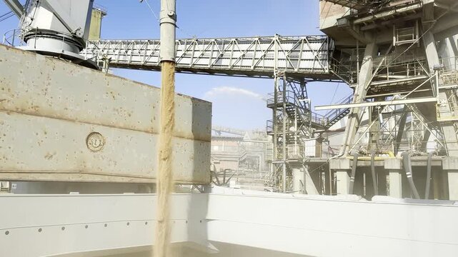 Heavy mechanical equipment transferring vast quantities of raw farming crop downward filling huge storage compartment aboard anchored aquatic freight carrier during sunny daytime dockyard operations.
