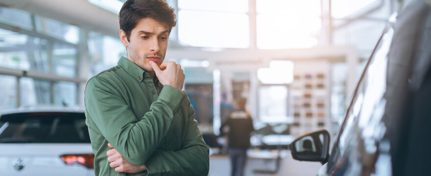 A man stands in a car dealership looking thoughtful while considering a vehicle. He appears focused and observant. The dealership has various cars displayed and is bright.