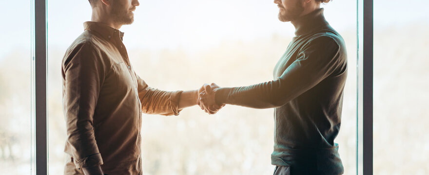 Two men stand facing each other and shake hands in a well-lit room with large windows. The background shows trees and nature, indicating a calm atmosphere.